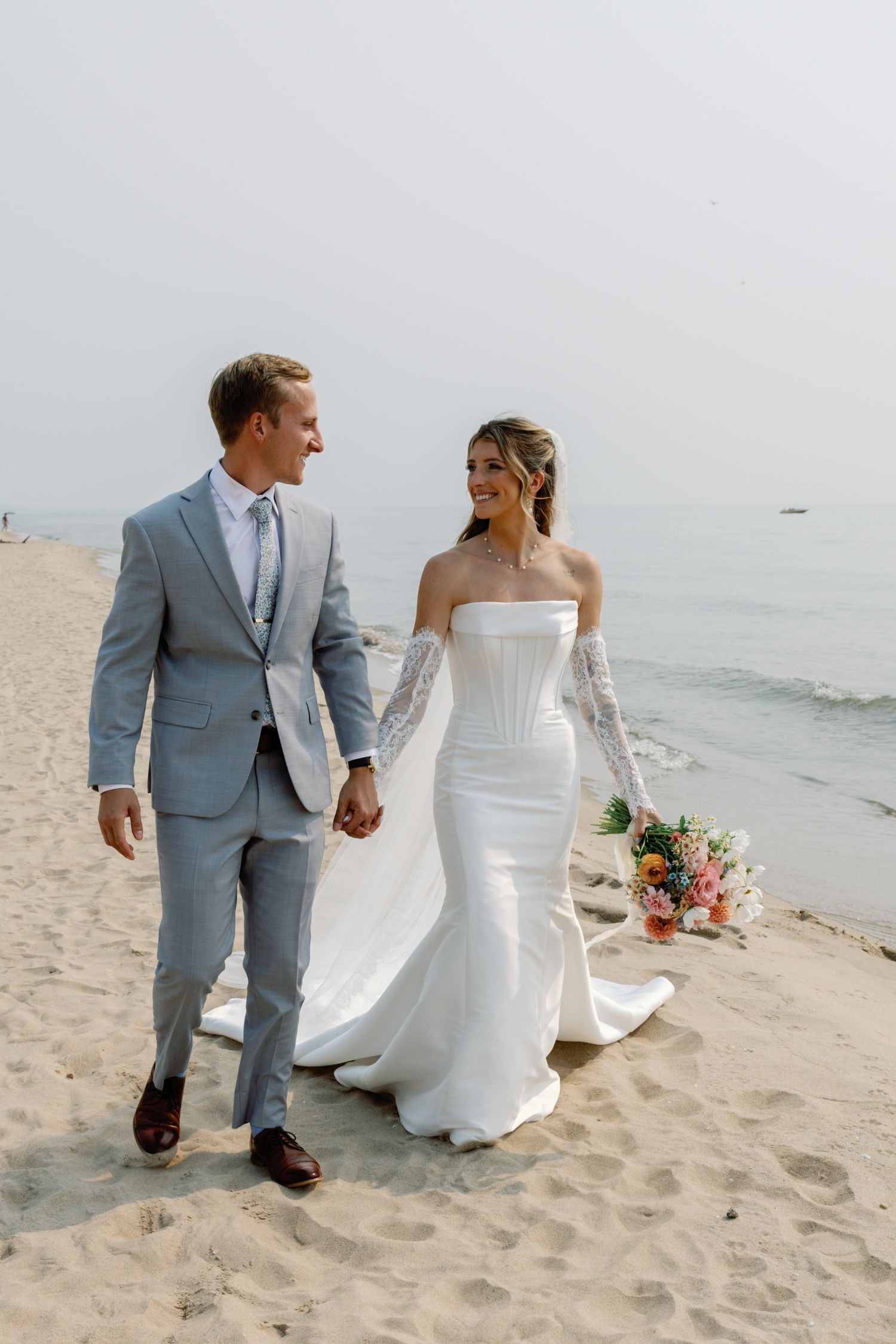 Newlyweds stroll along the shoreline with bride's dress and veil flowing in the wind.