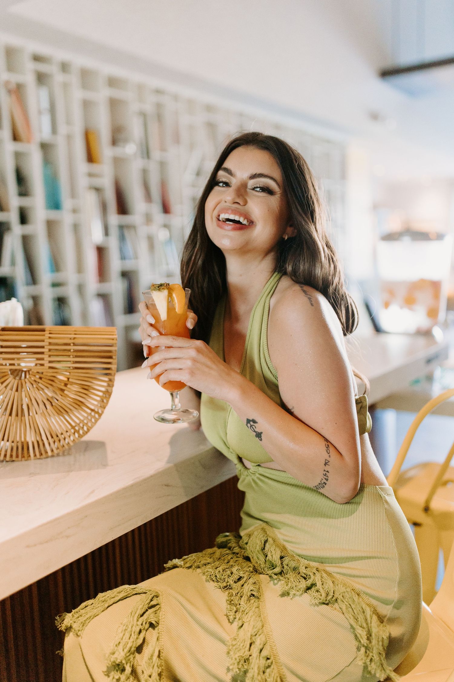 Woman in green dress laughing while holding an orange cocktail at a bar with wicker decor.