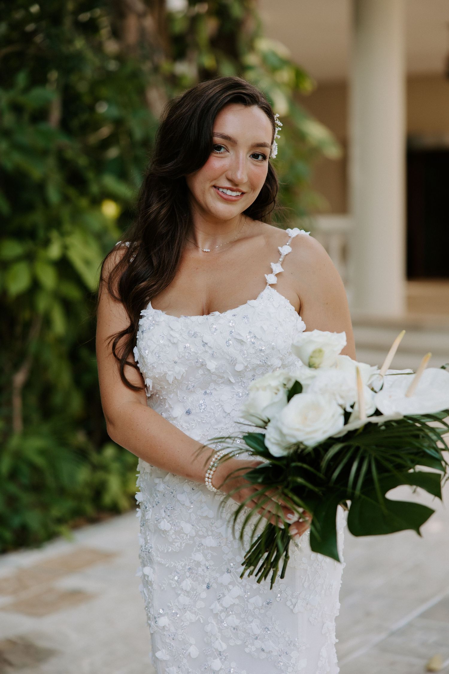 Bride in sparkling white dress holding modern tropical bouquet with white flowers and monstera leaves.