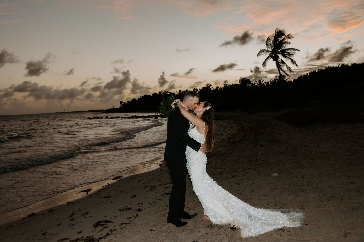Silhouetted wedding couple embracing on a beach at sunset with palm trees in the background.