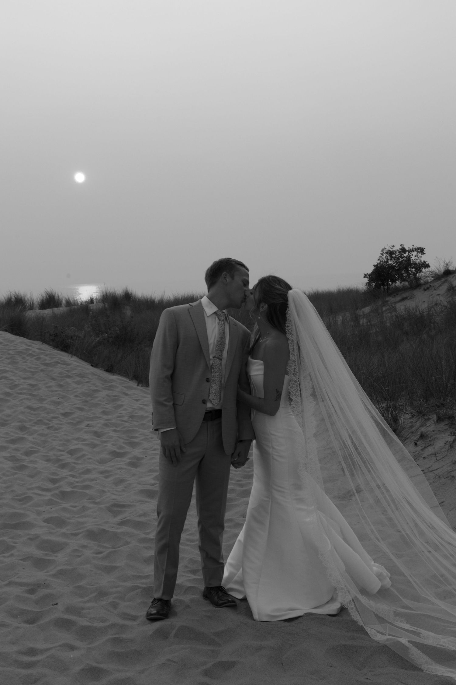 Black and white wedding photos of a couple on a beach with flowing veil and sand dunes.