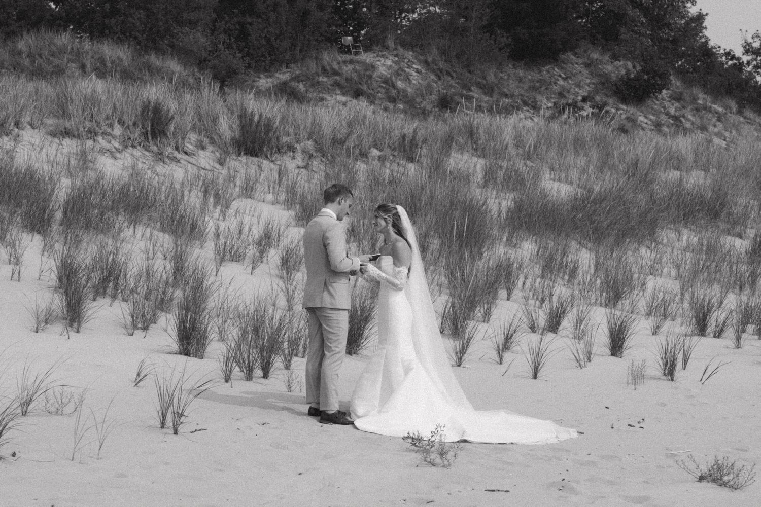 Long white wedding dress train extends across sandy beach dunes with grass.