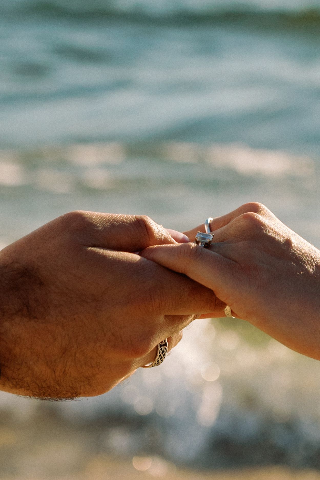 Close-up of hands with engagement rings against sparkling ocean water background.