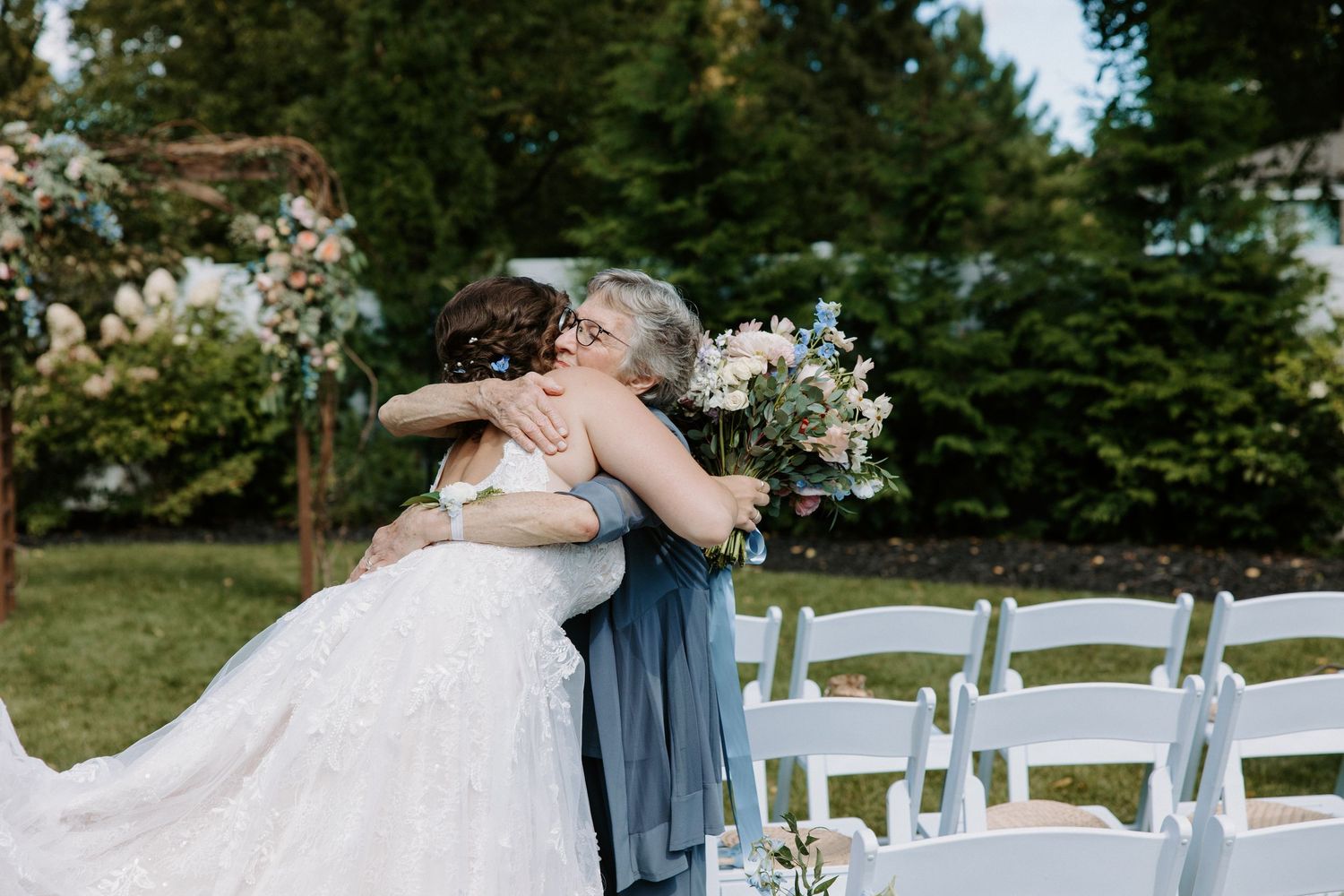 Outdoor wedding ceremony setup with white chairs and floral arrangements in natural garden setting.