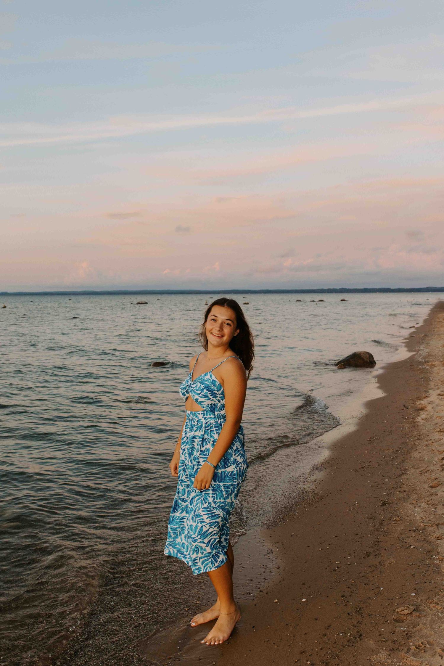 Figure in blue patterned dress stands barefoot on rocky beach during evening light.