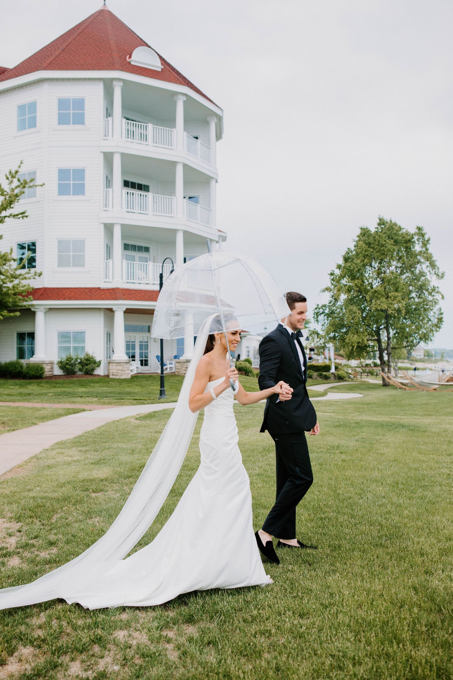 A bride and groom walk together near a coastal mansion, with the bride holding a clear umbrella.