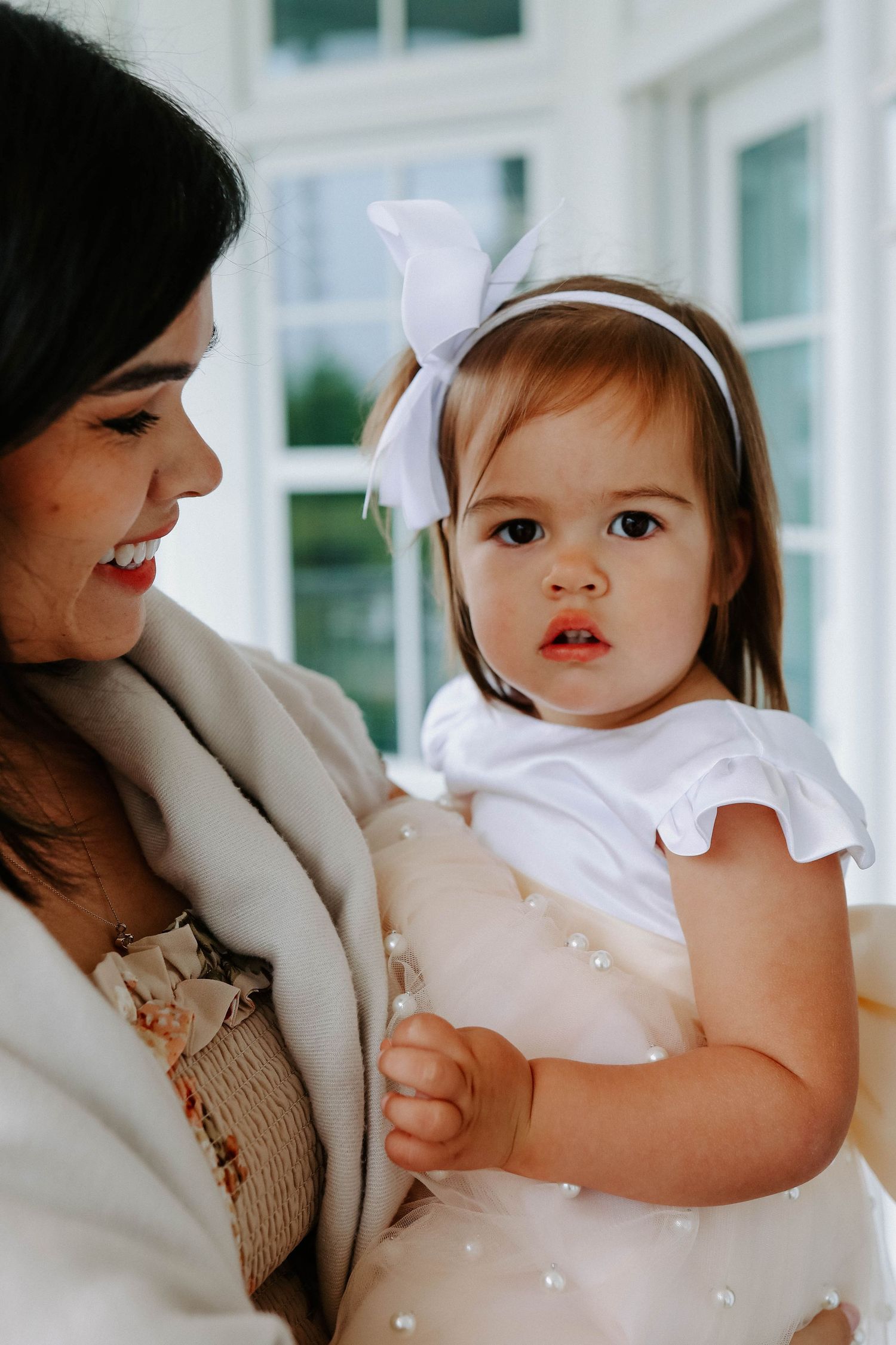 A tender moment captured between a woman in beige and a baby wearing a white dress with a bow headband.