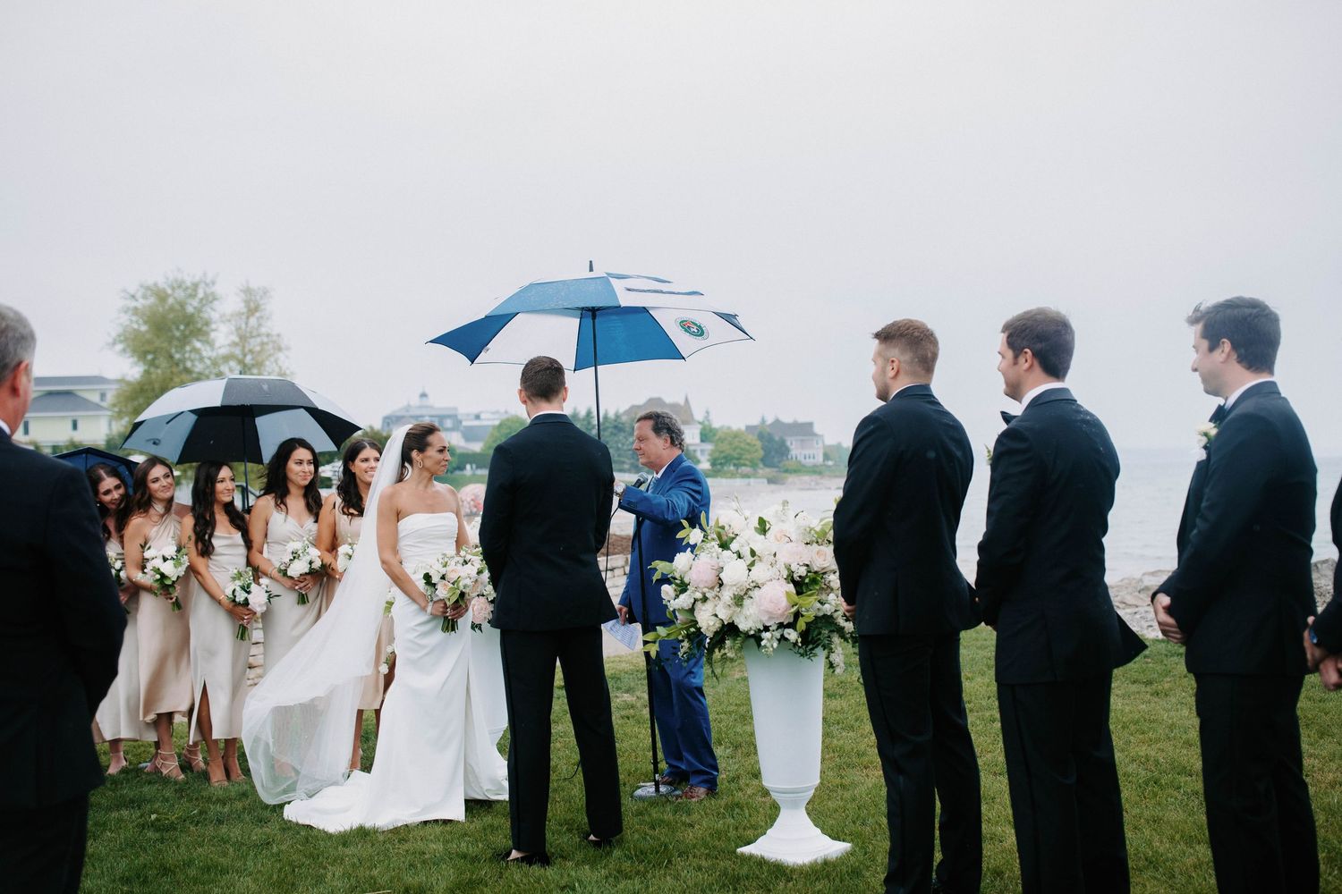 Wedding party members gather outdoors on grass during an overcast ceremony with umbrellas.
