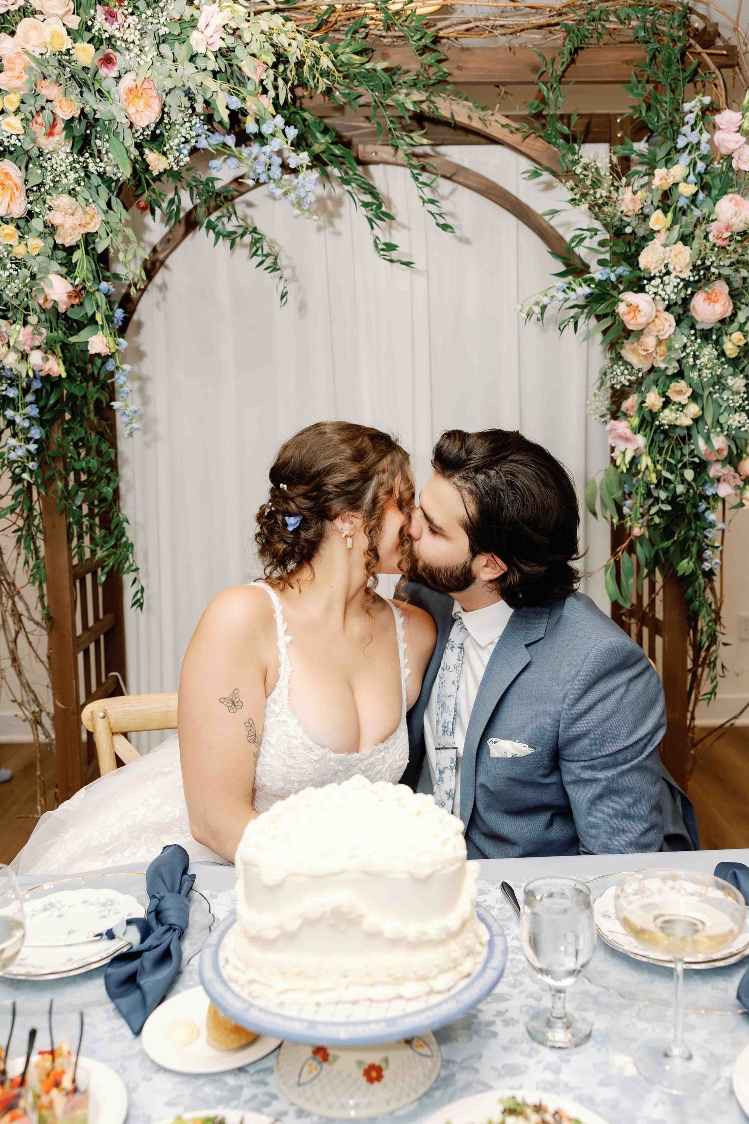A couple shares an intimate moment at their wedding reception table decorated with floral arrangements and a white cake.