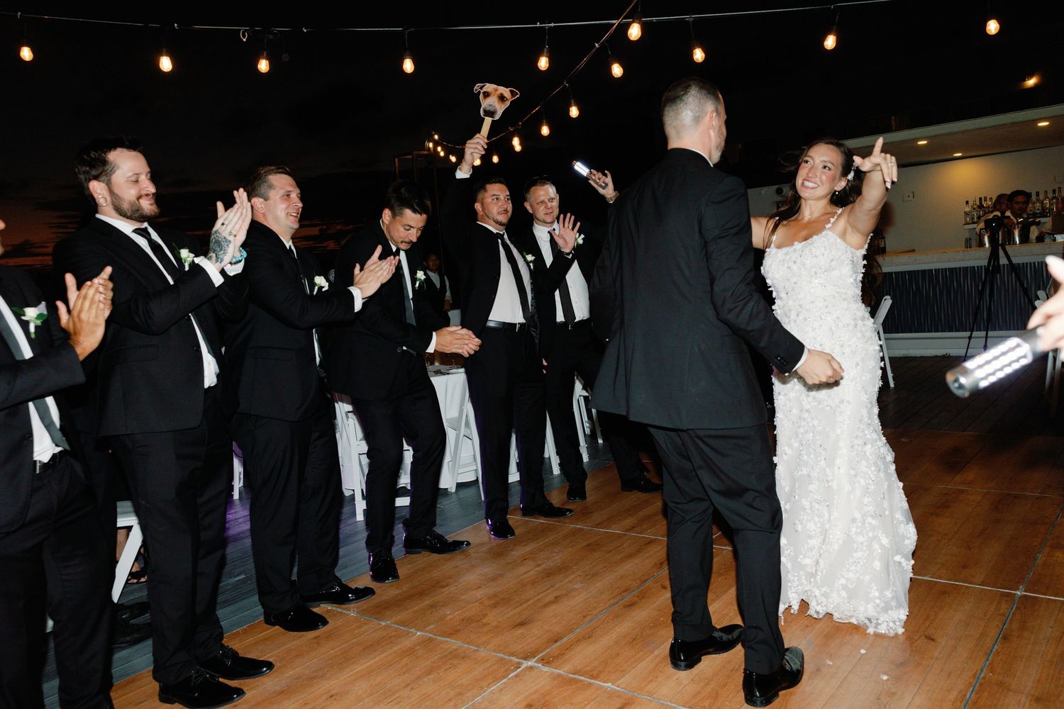 Wedding guests in black suits cheer and clap as a couple dances during a reception celebration.