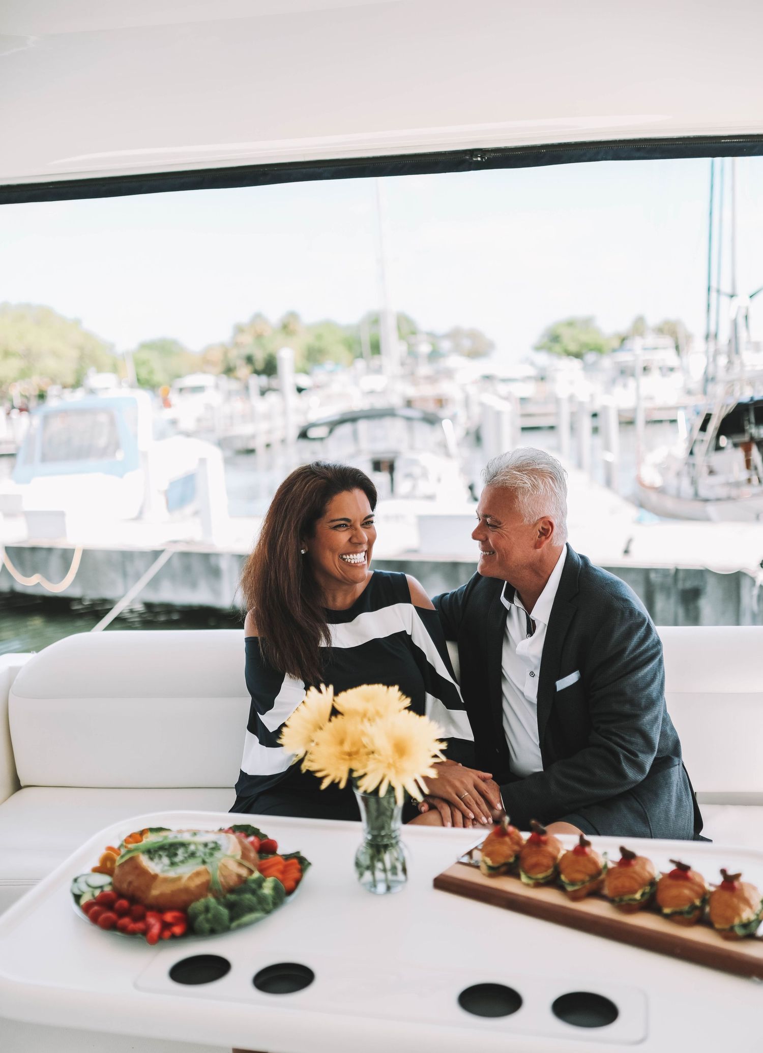 A couple enjoys an intimate dinner at a waterfront restaurant with flowers, appetizers and a marina view in the background.