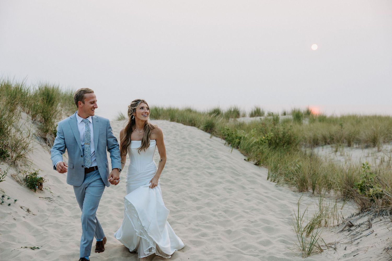 Newlyweds walking together along a sandy beach path with coastal vegetation.