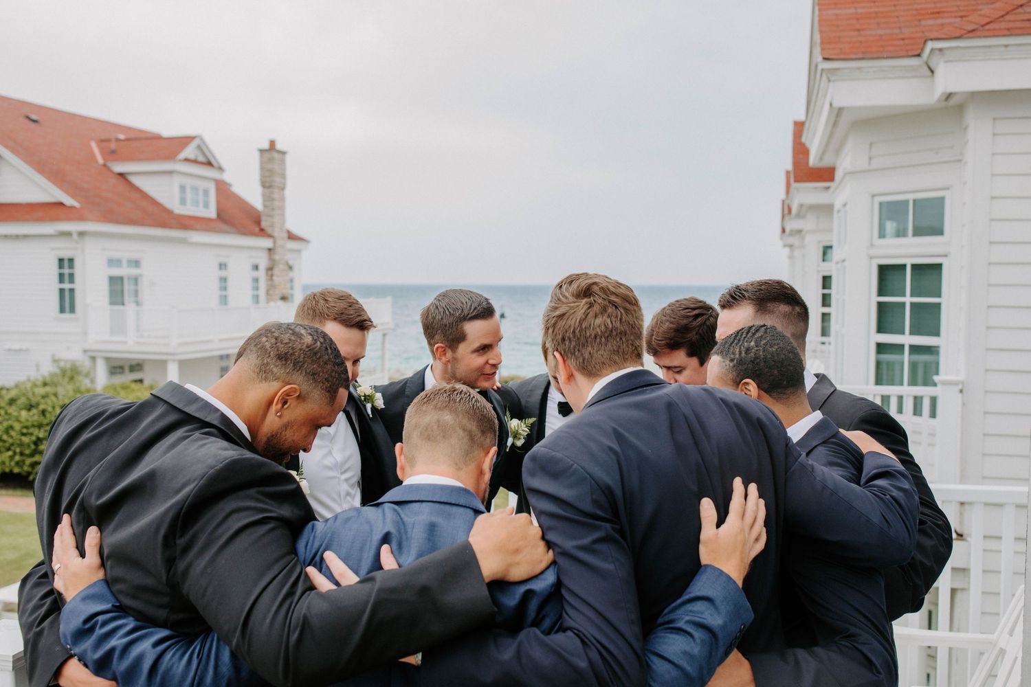 Groomsmen in formal attire embrace in a group hug on a coastal porch.