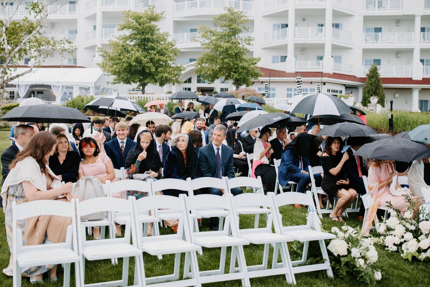 Wedding guests seated in white chairs at outdoor ceremony with hotel building in background.