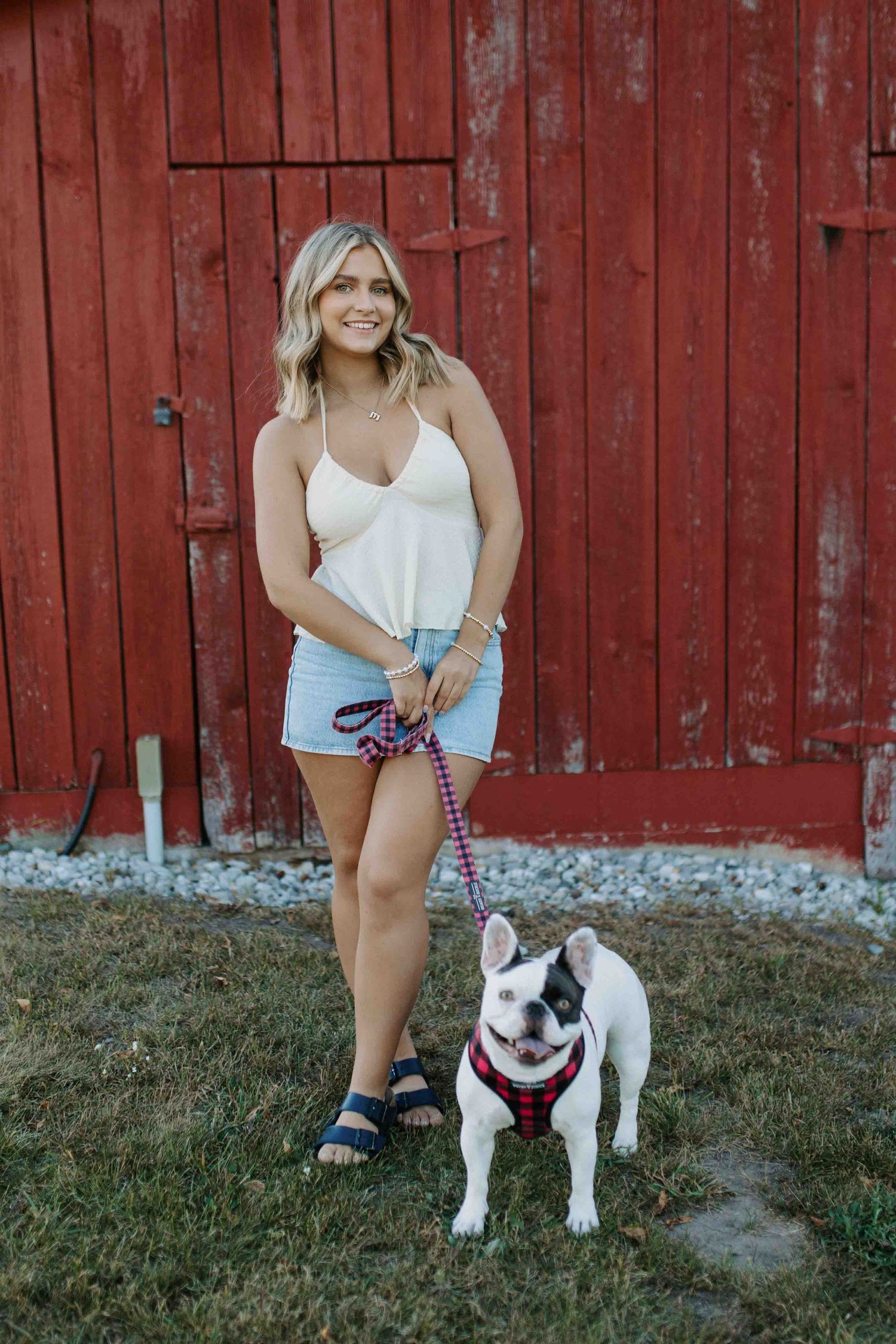Woman in summer outfit poses with happy French Bulldog by red barn.