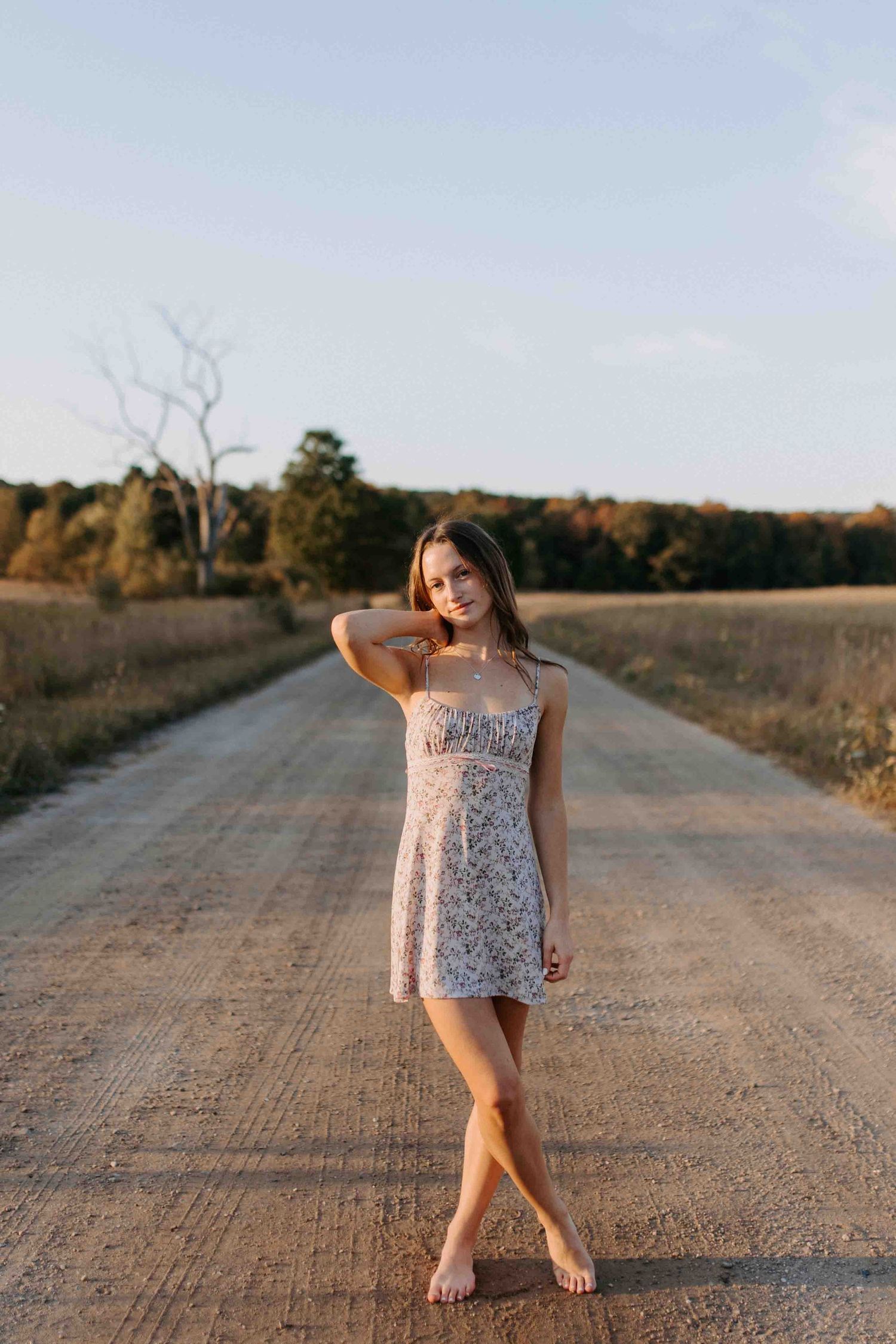 Someone in a floral dress poses playfully on a dirt road during golden hour.