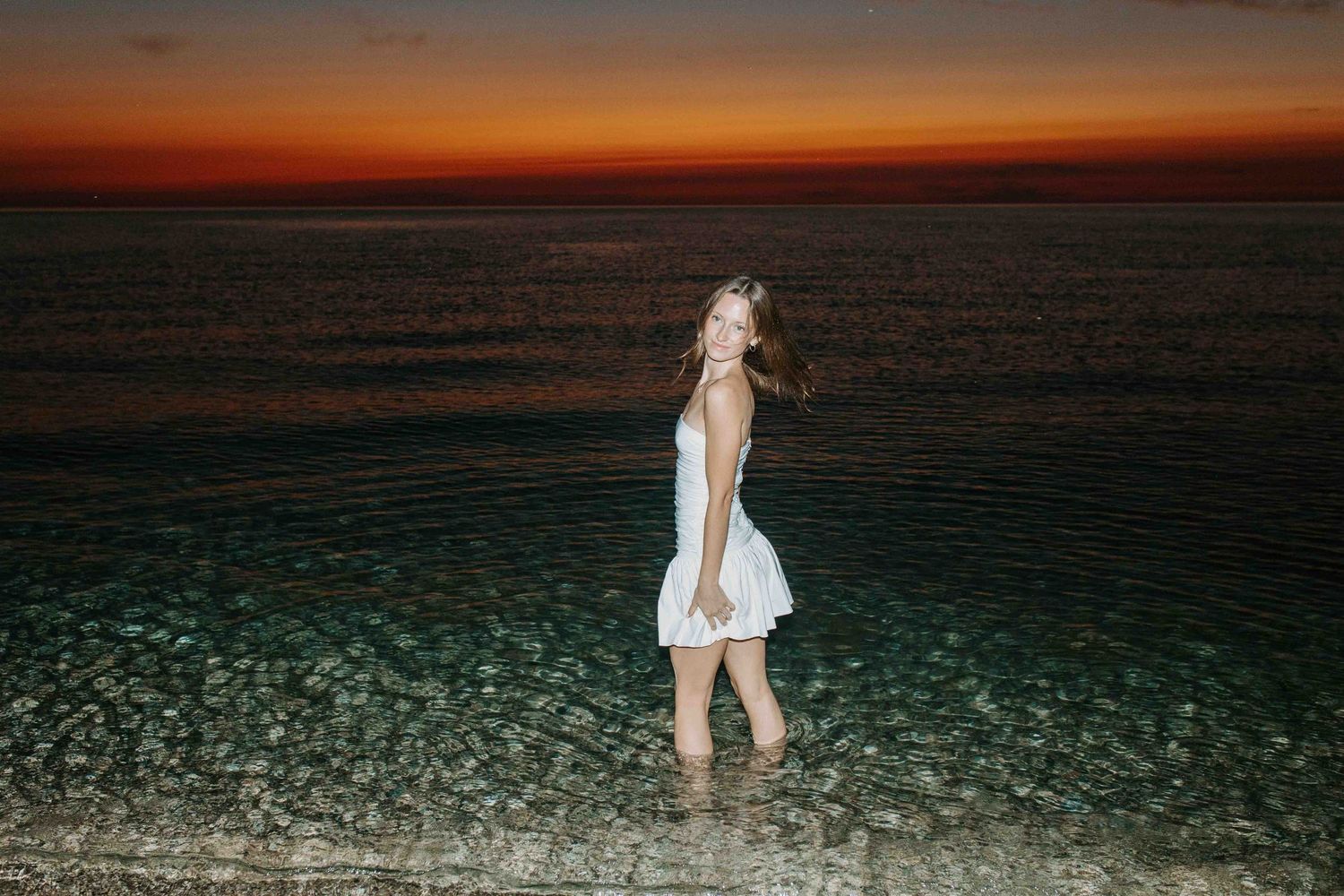 People playing in shallow ocean water at sunset with dramatic orange sky.