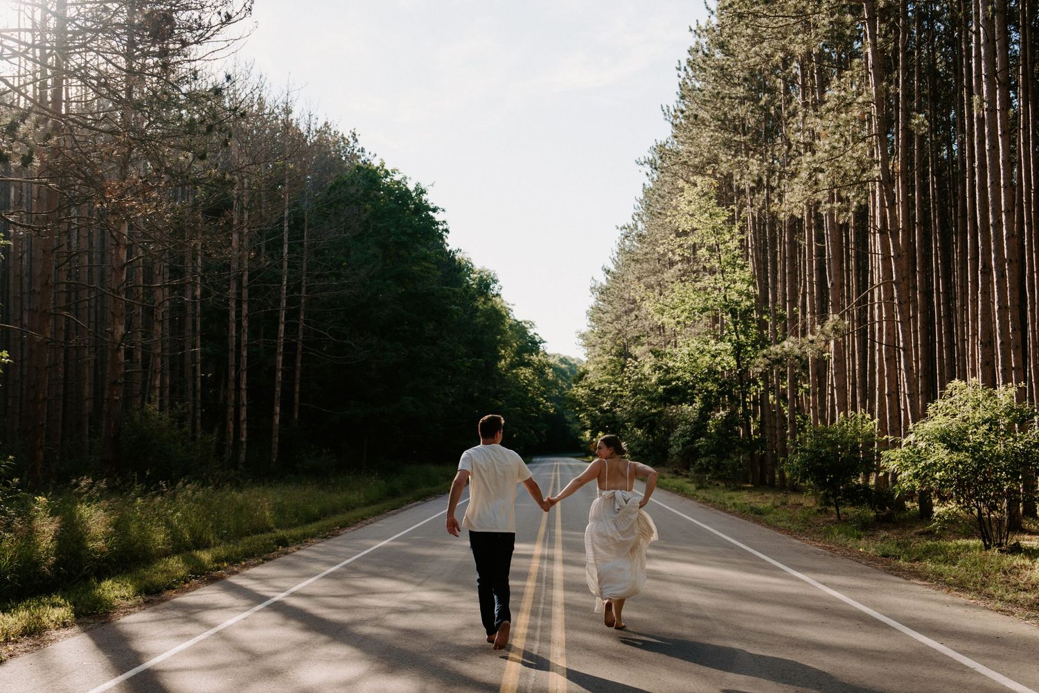 A couple walks down a long tree-lined road through a dense forest.