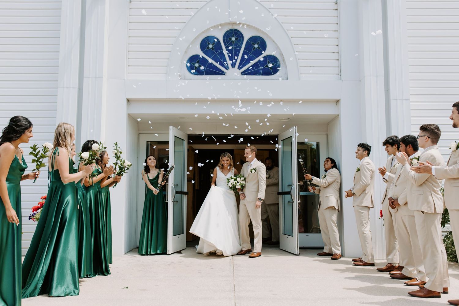 Wedding party in emerald green dresses and tan suits lined up in front of white building with string lights.