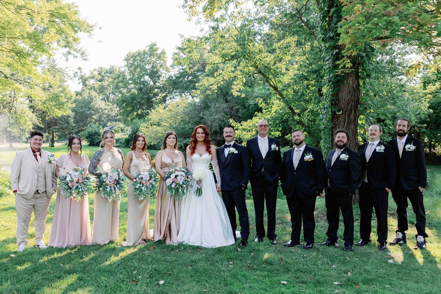 Wedding party in formal attire stands in line outdoors with trees in background.