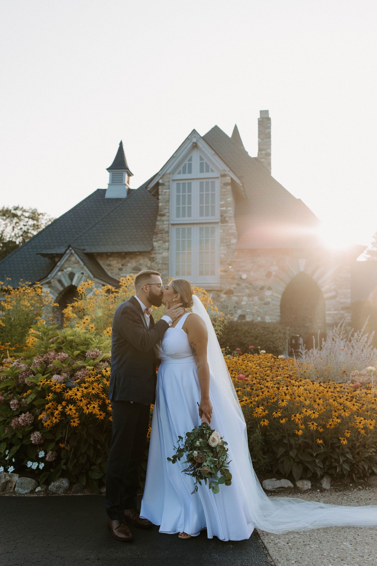 Wedding couple poses in front of stone mansion with golden wildflowers in foreground at sunset.