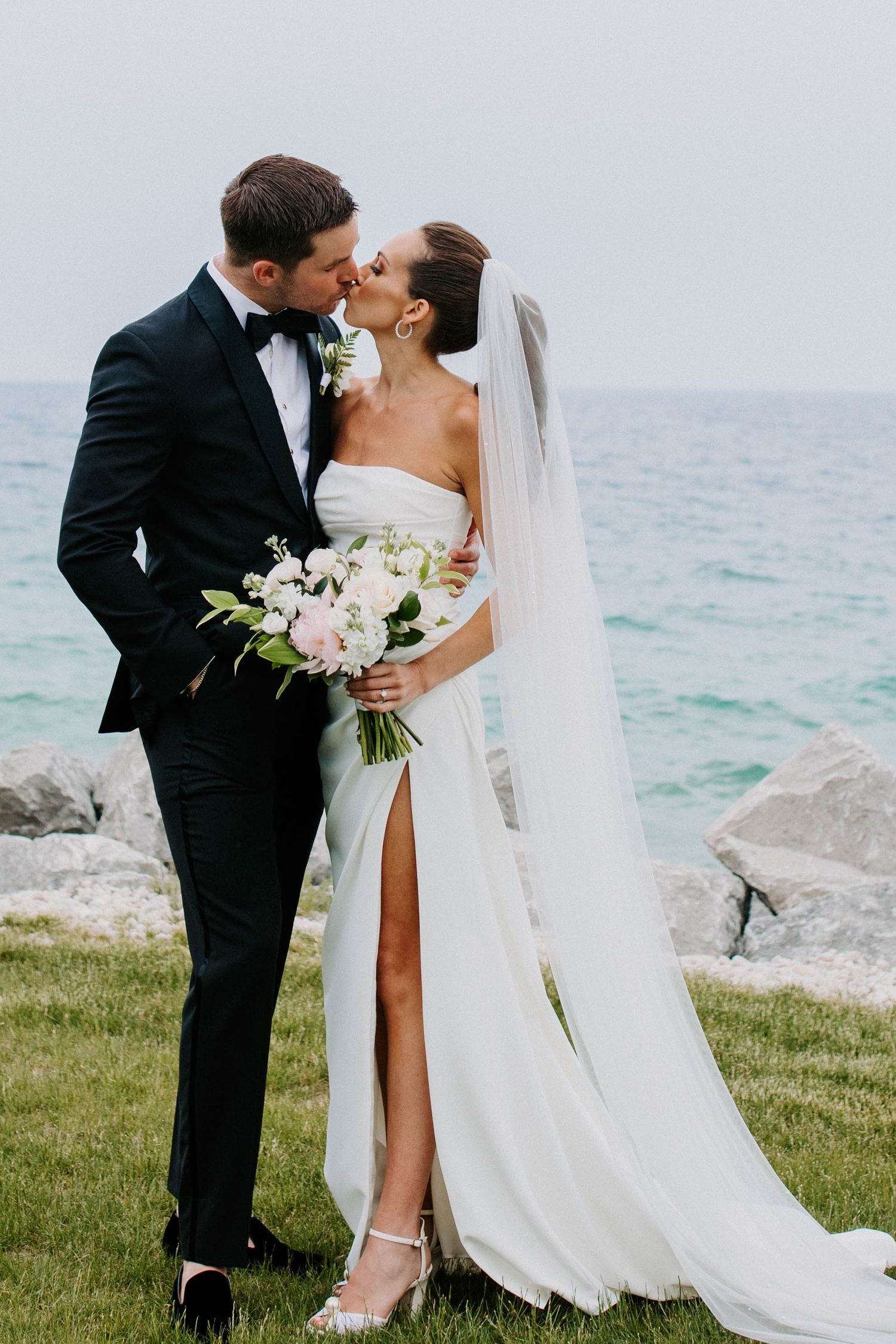 Wedding couple poses romantically by the ocean with flowing white dress and bouquet.