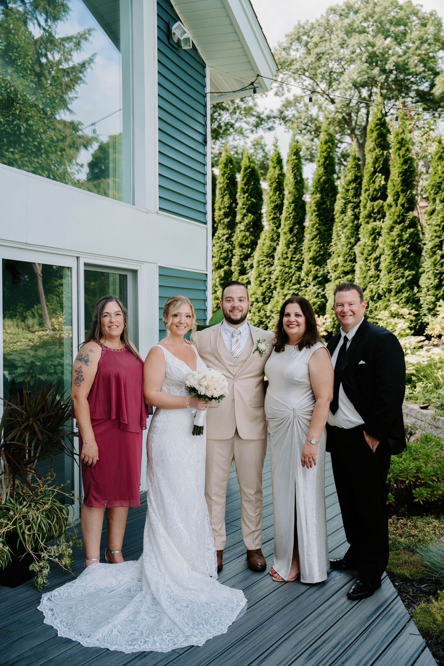 Wedding party poses together in front of a white house with green siding and evergreen trees.