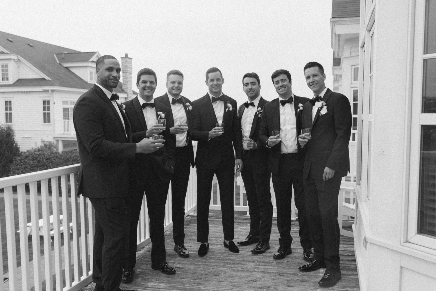 Groomsmen lined up in black tuxedos on a white coastal porch.
