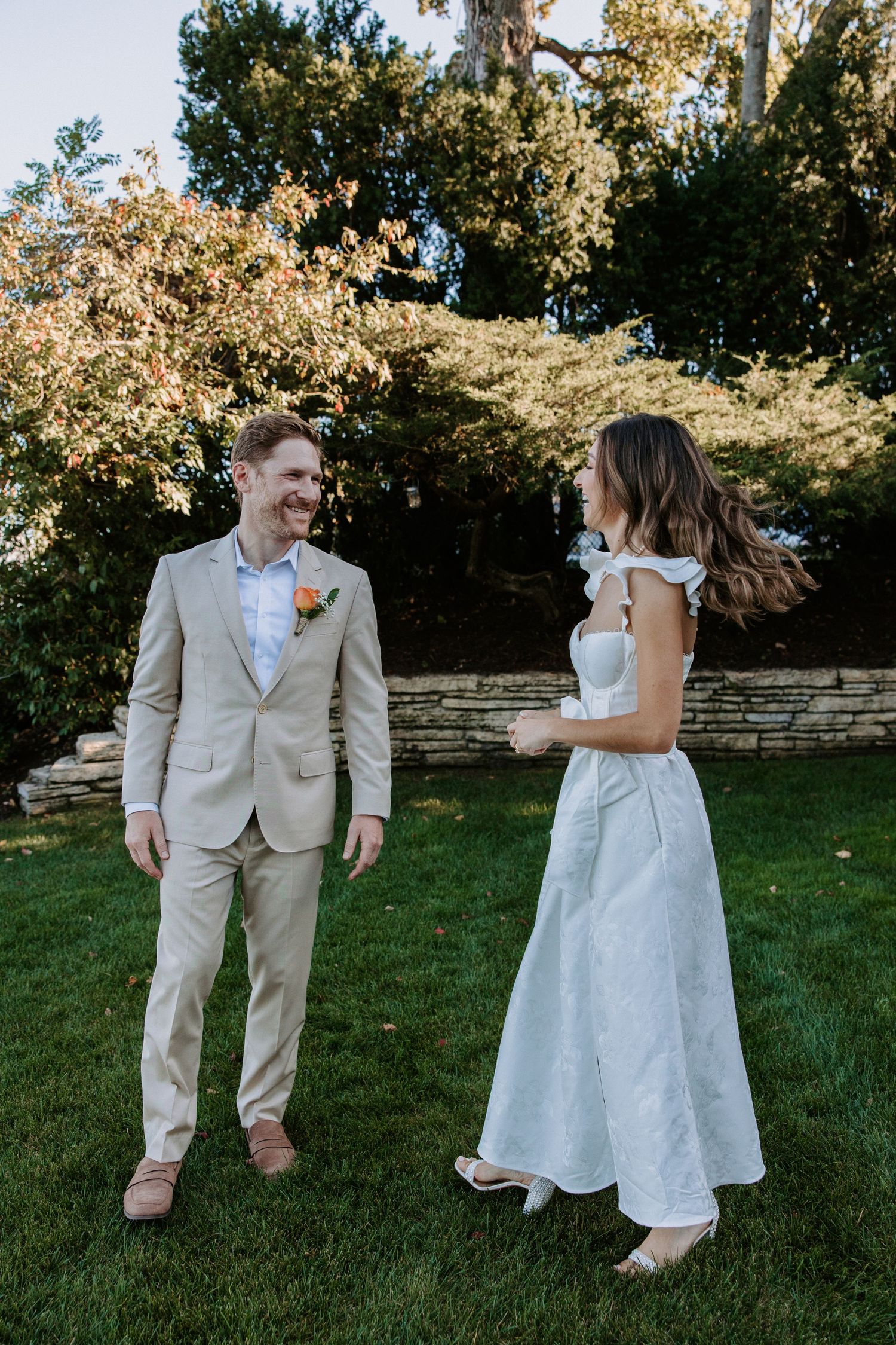 Couple in wedding attire dancing playfully on grass with stone wall and trees in background.