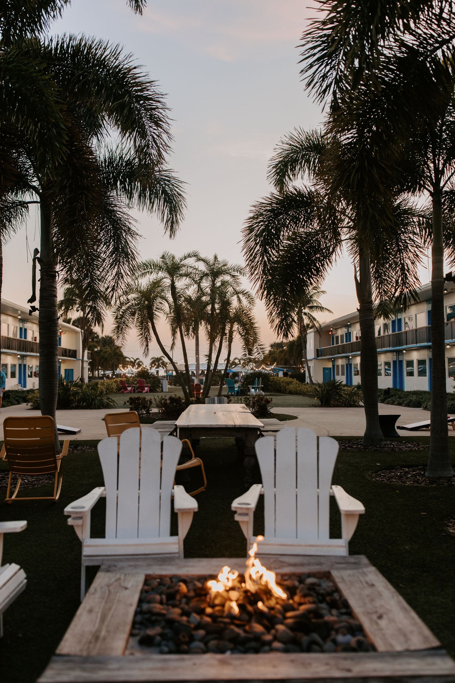 Two white Adirondack chairs face a fire pit at dusk with palm trees and coastal buildings in the background.