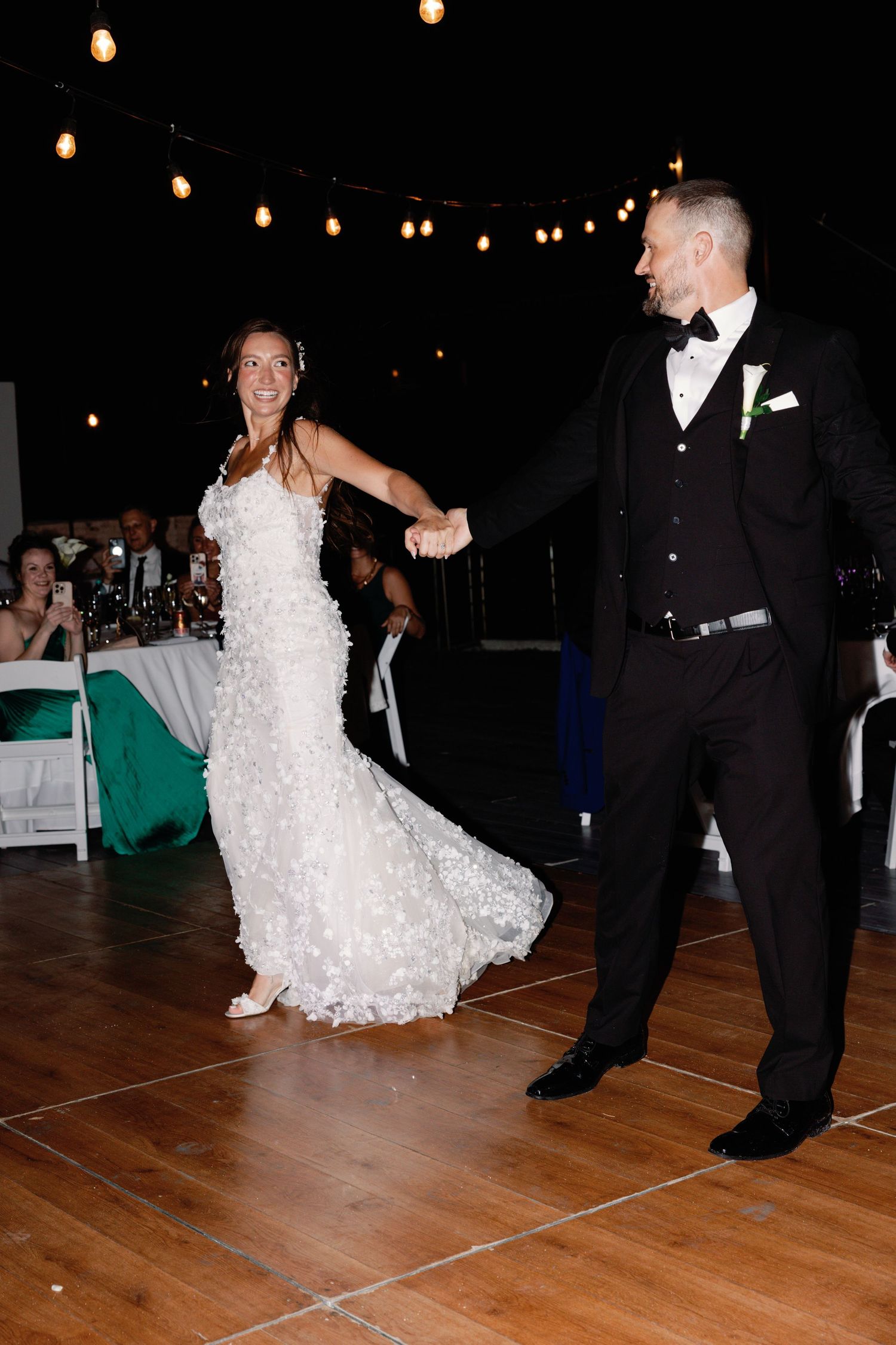 Newlyweds dance together on wooden floor under string lights at reception venue.