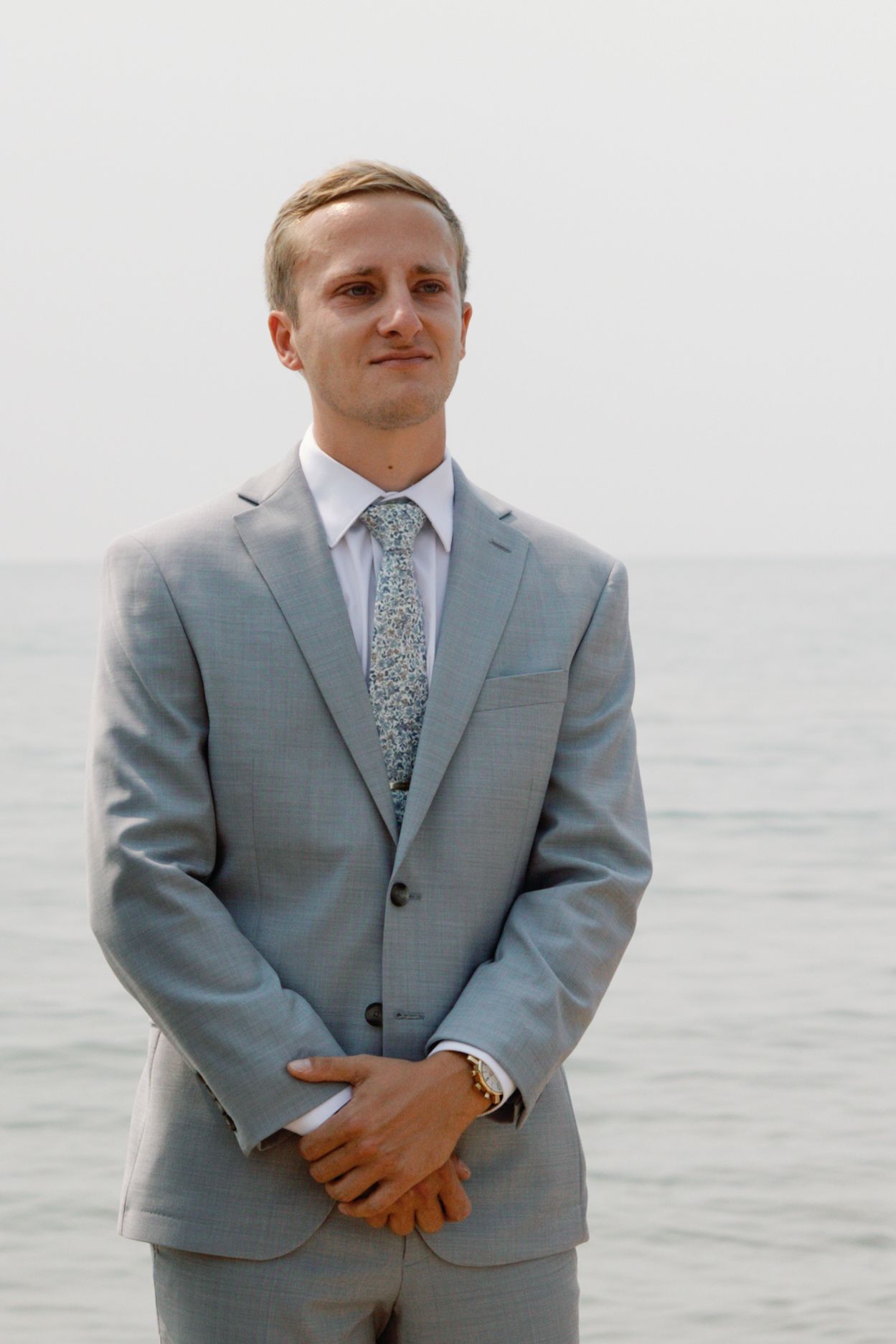 Groom in light gray suit standing by the ocean on his wedding day.