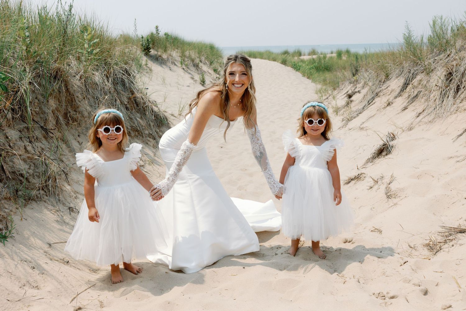 Bridesmaids in light colored dresses gather for photos on sand dunes at the beach.