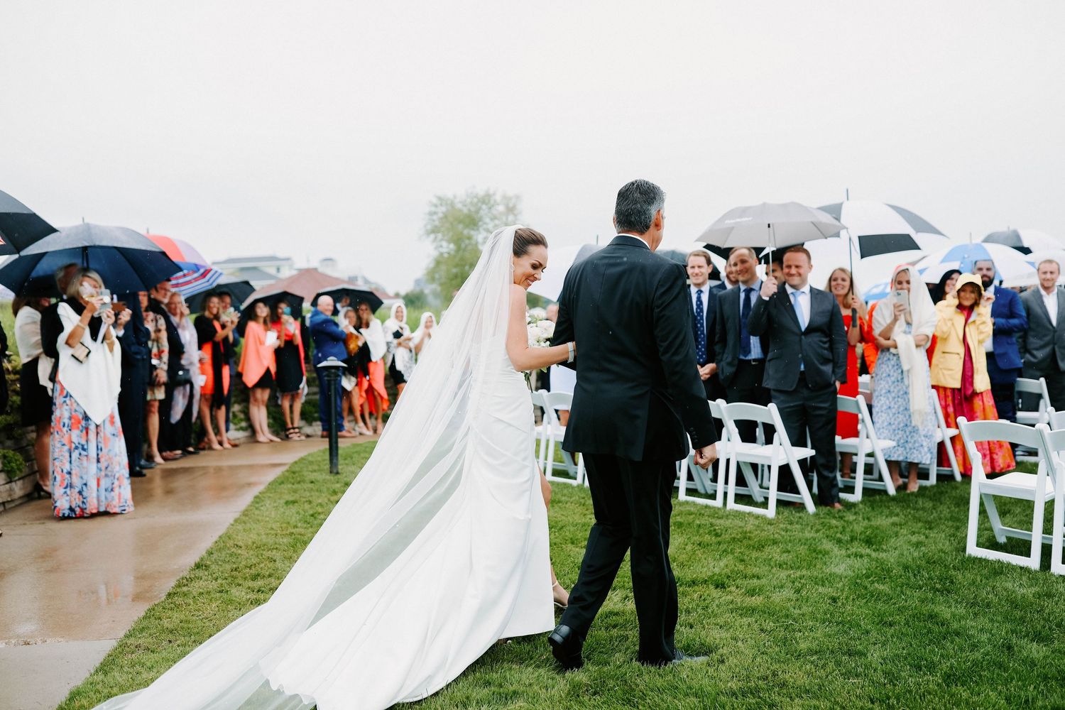 Outdoor wedding ceremony with guests holding umbrellas on a cloudy day by the water.