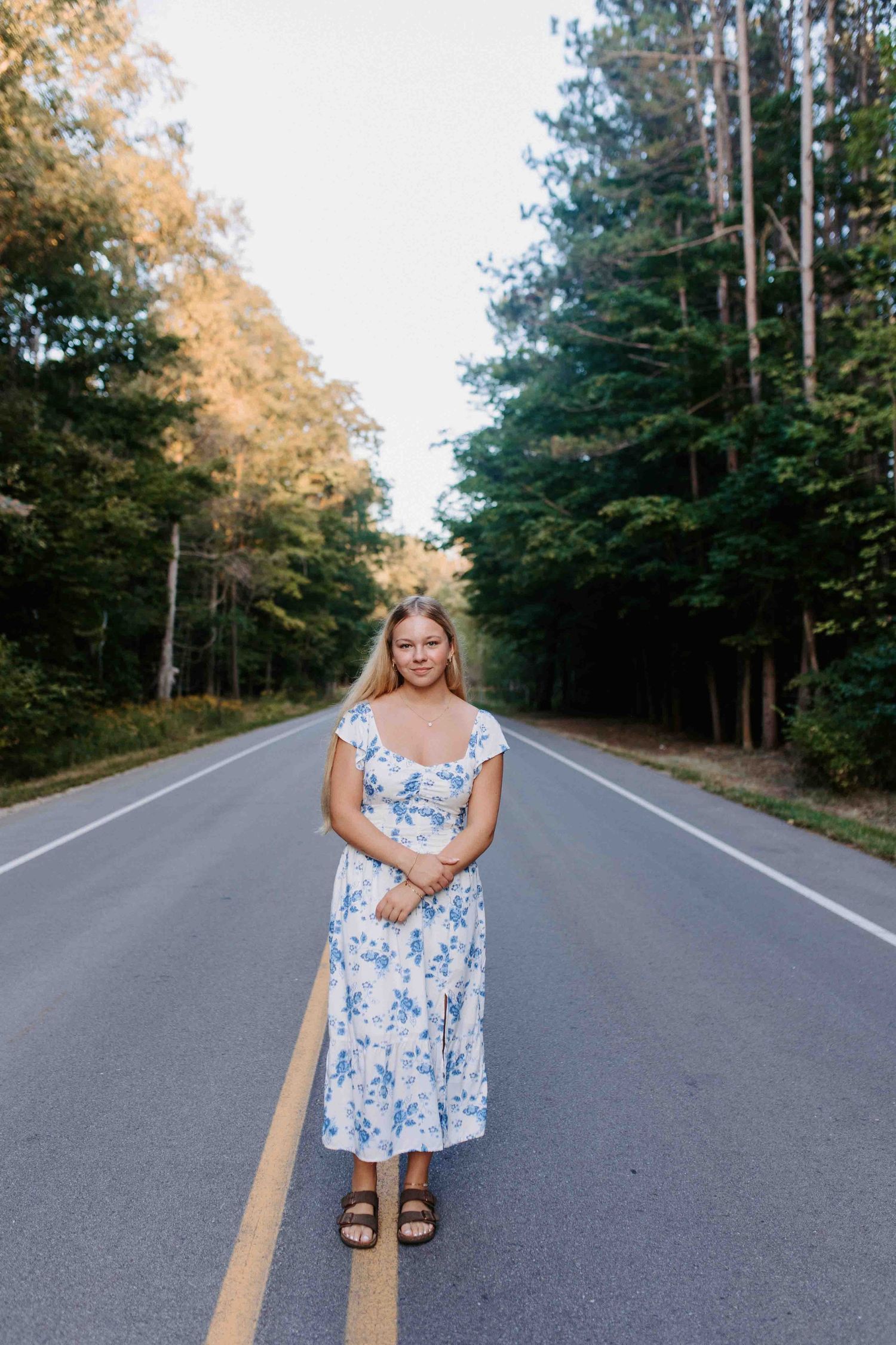 Person in a blue floral dress poses playfully on a tree-lined road on a sunny day.