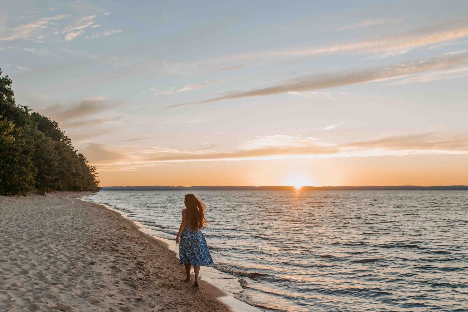 A figure in a blue dress walks along a sandy beach at sunset with calm waters and pink sky in the background.