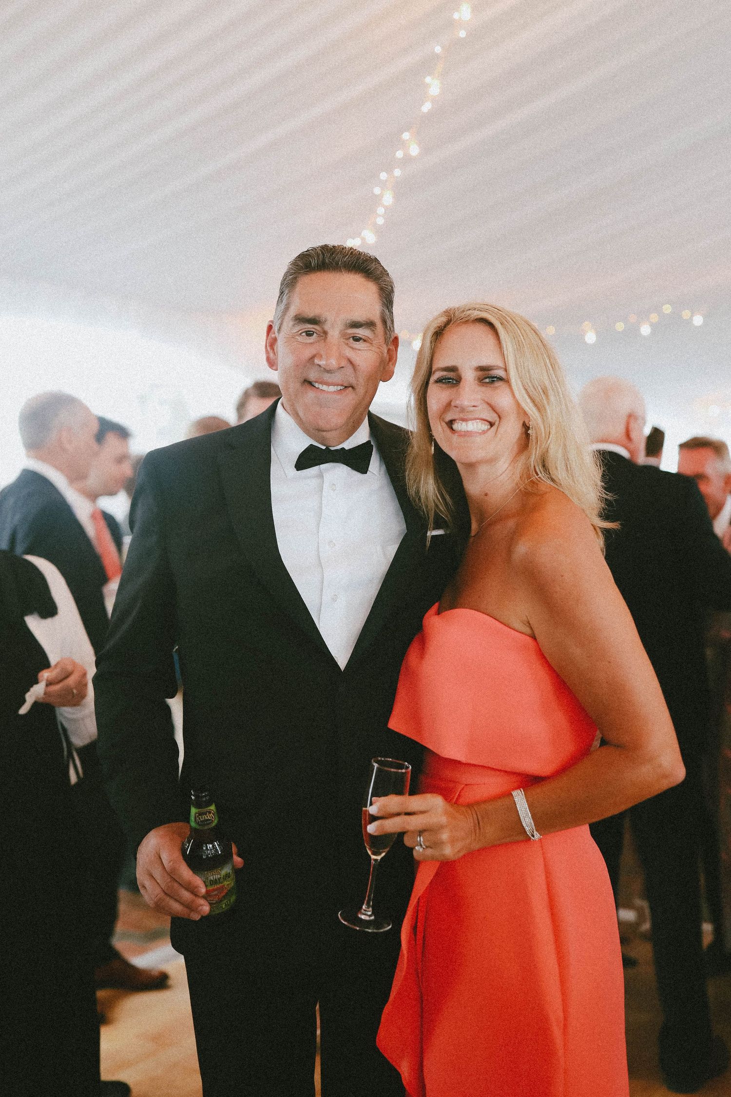 A couple in formal attire, with the woman wearing a vibrant coral dress, share a joyful moment at an elegant event.