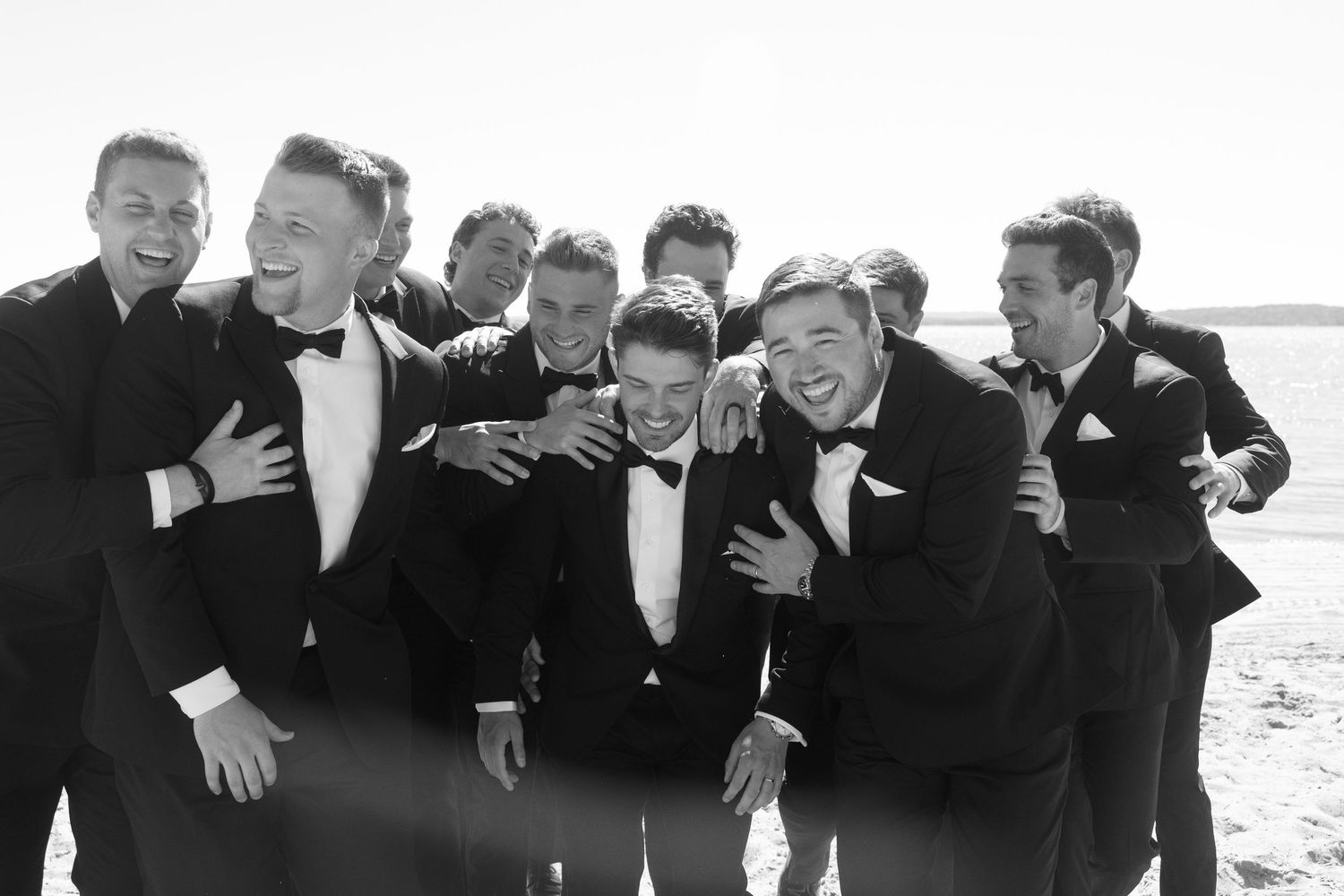 Wedding party in black attire poses playfully on the beach for group photos.
