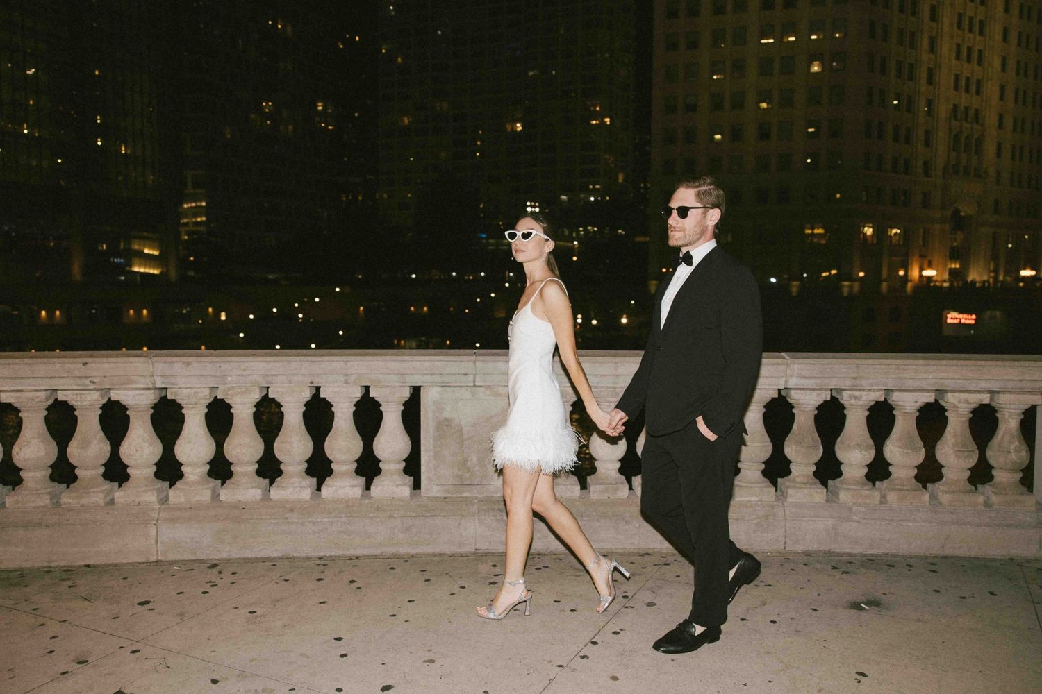 Couple walking along an elegant balcony at night with city lights in the background.