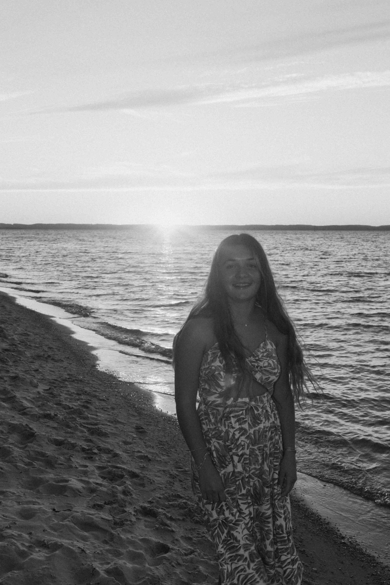 Black and white photo of person in patterned dress on beach at sunset.