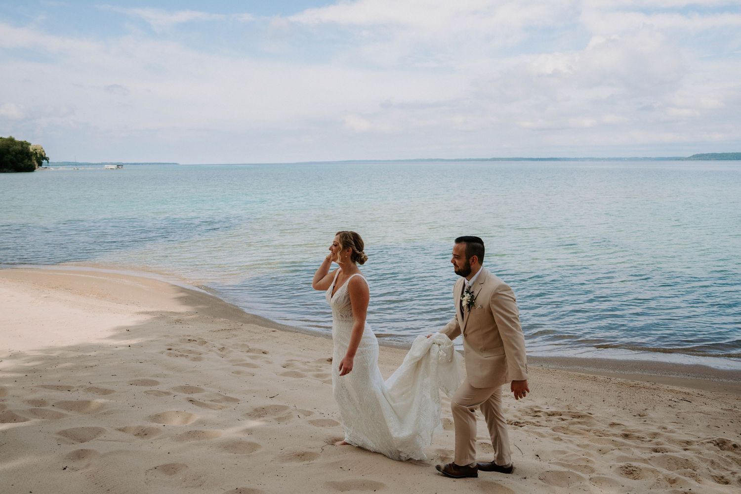 Newlyweds walk hand in hand along a sandy beach with calm ocean waters stretching to the horizon.