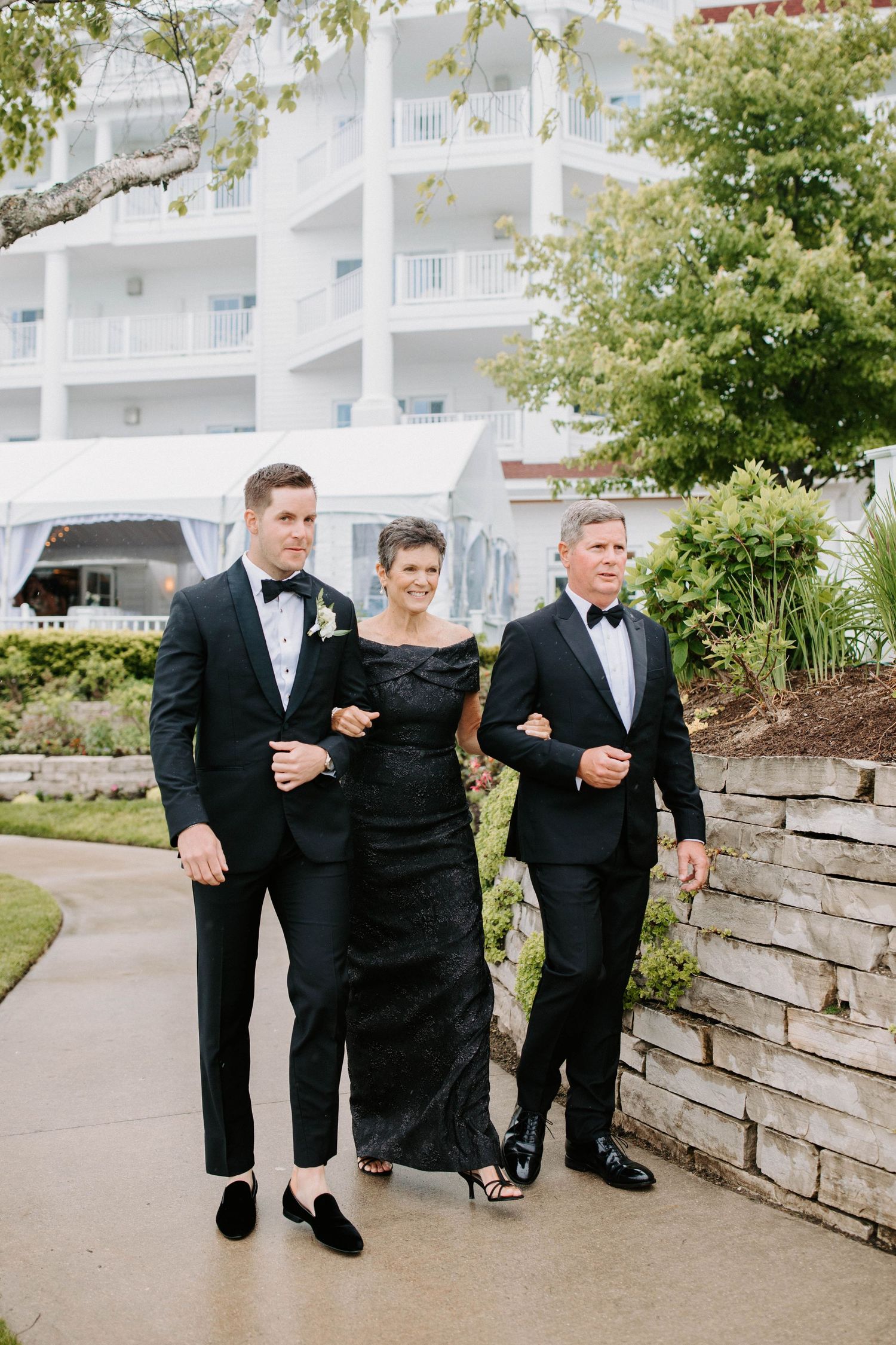 Wedding party in formal attire walks together along a stone retaining wall pathway.