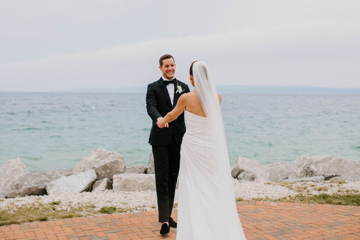 A wedding couple embraces on a brick patio overlooking the ocean.