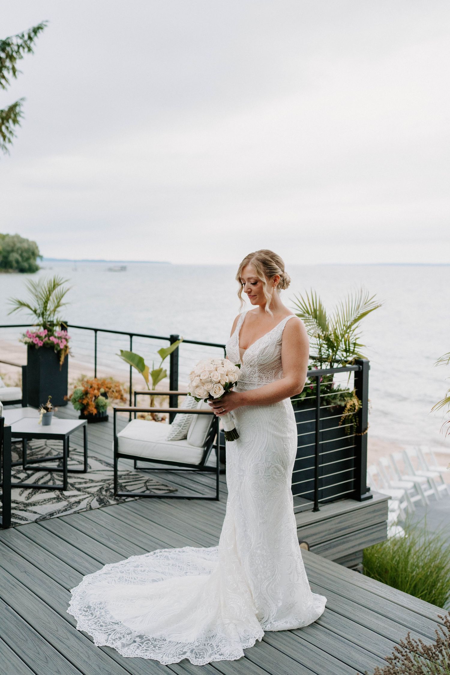 Bride in lace wedding dress poses on oceanfront deck with bouquet.