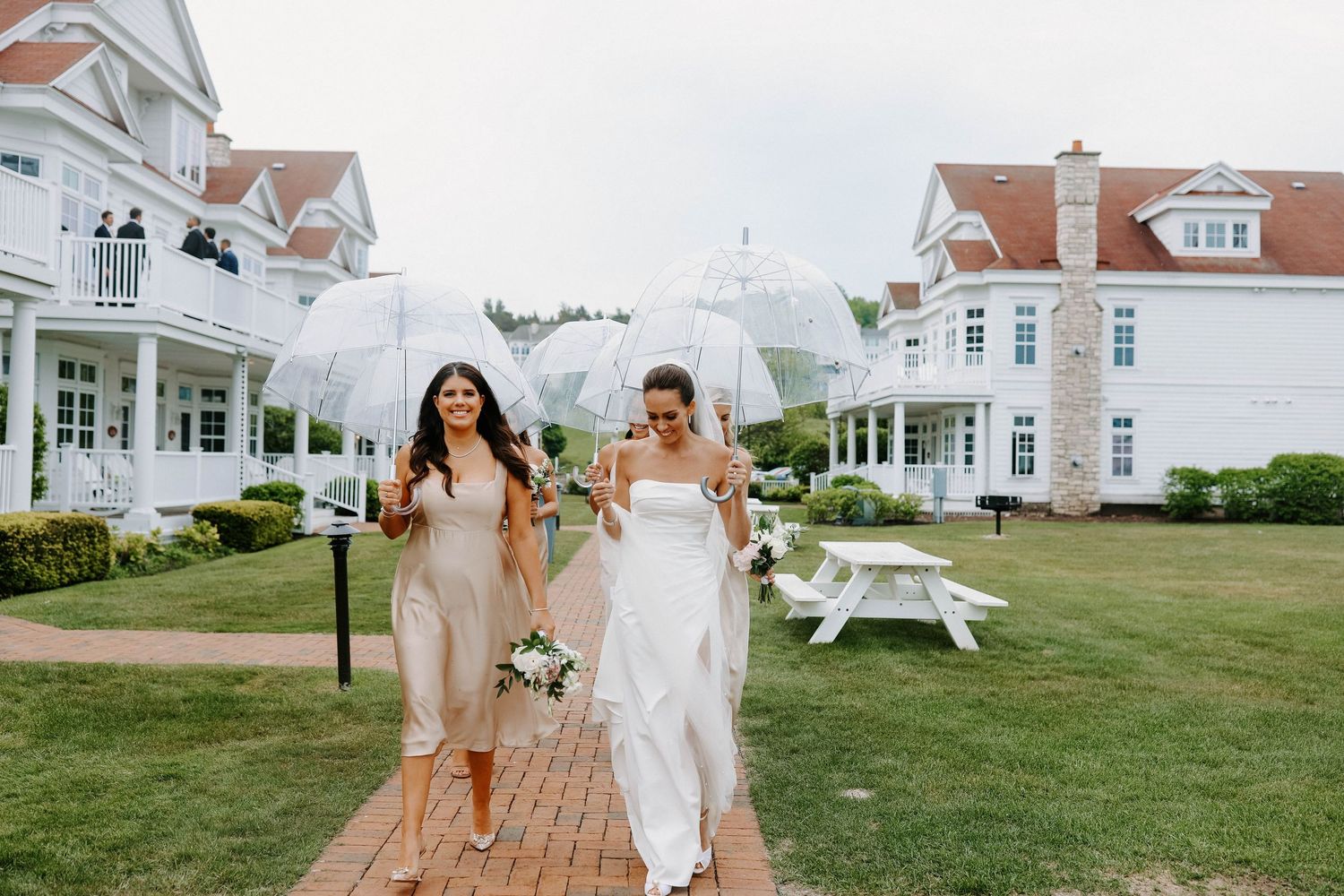 Two people walking down a brick path with white umbrellas near coastal houses.