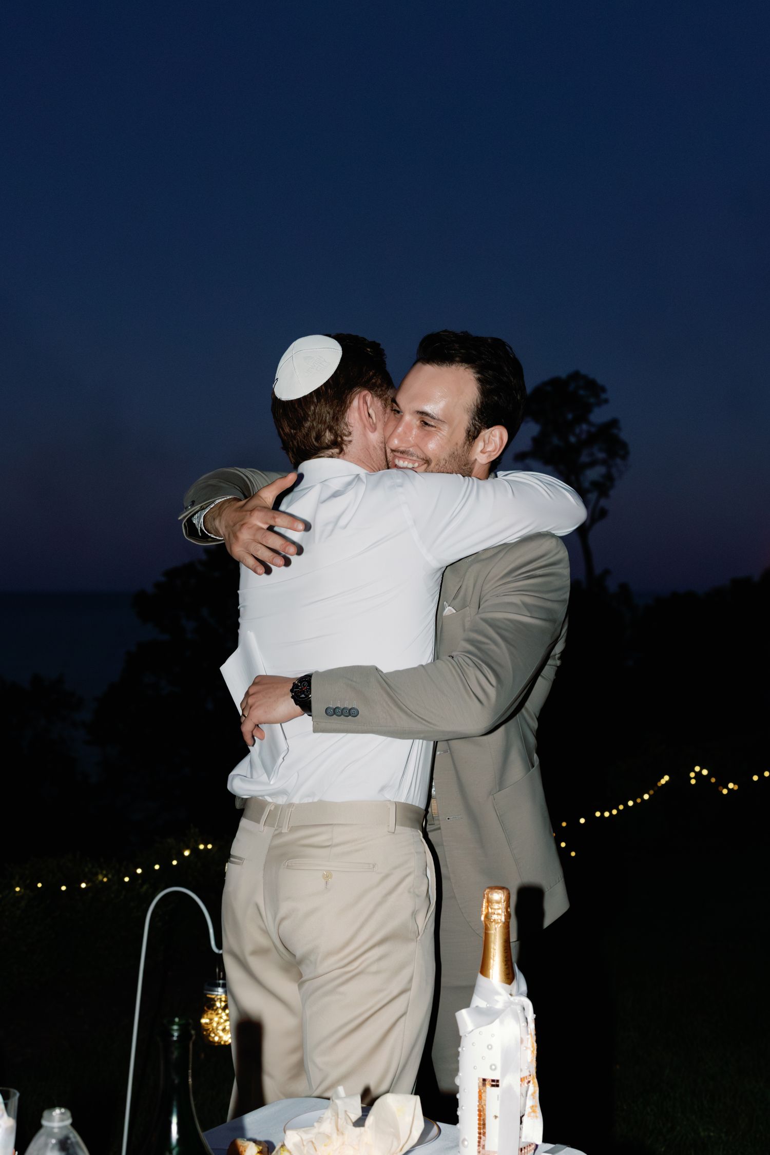Emotional embraces between people at a nighttime outdoor celebration with string lights.