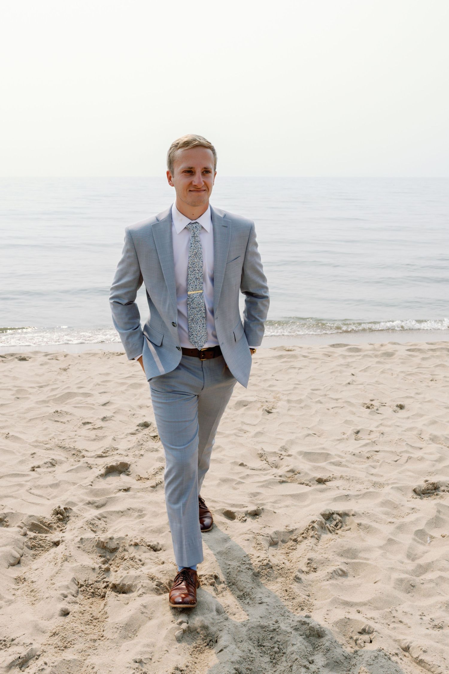 Groom in light blue suit walking along sandy beach shoreline.