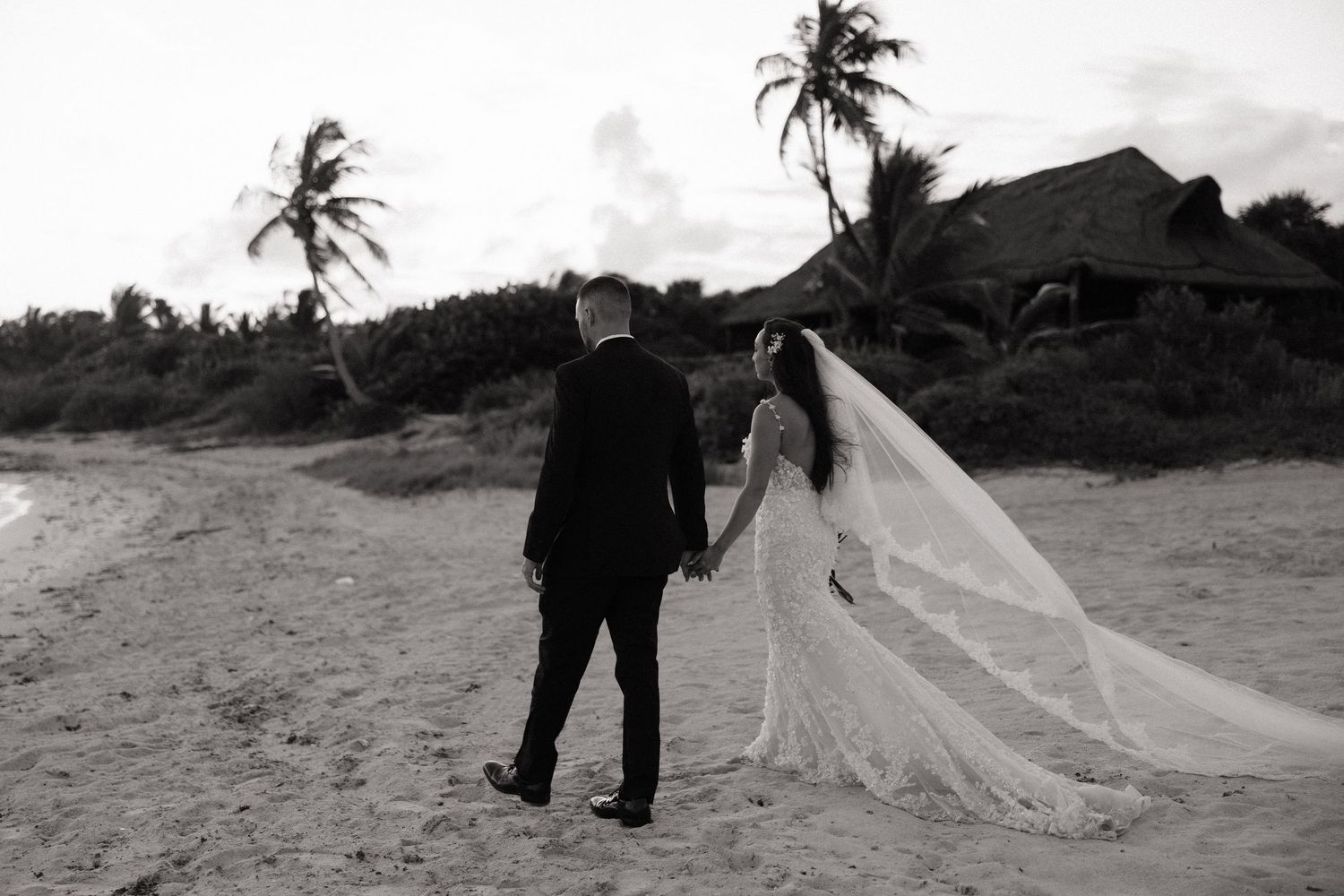 A couple stands on a beach with palm trees and a flowing veil in black and white.