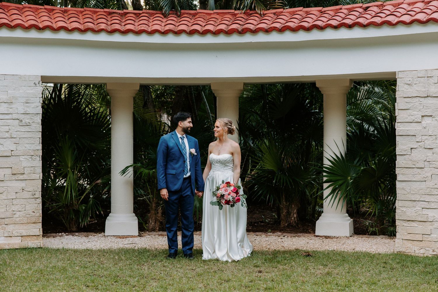 A couple stands together under a white stone gazebo with a terracotta roof and tropical foliage in the background.