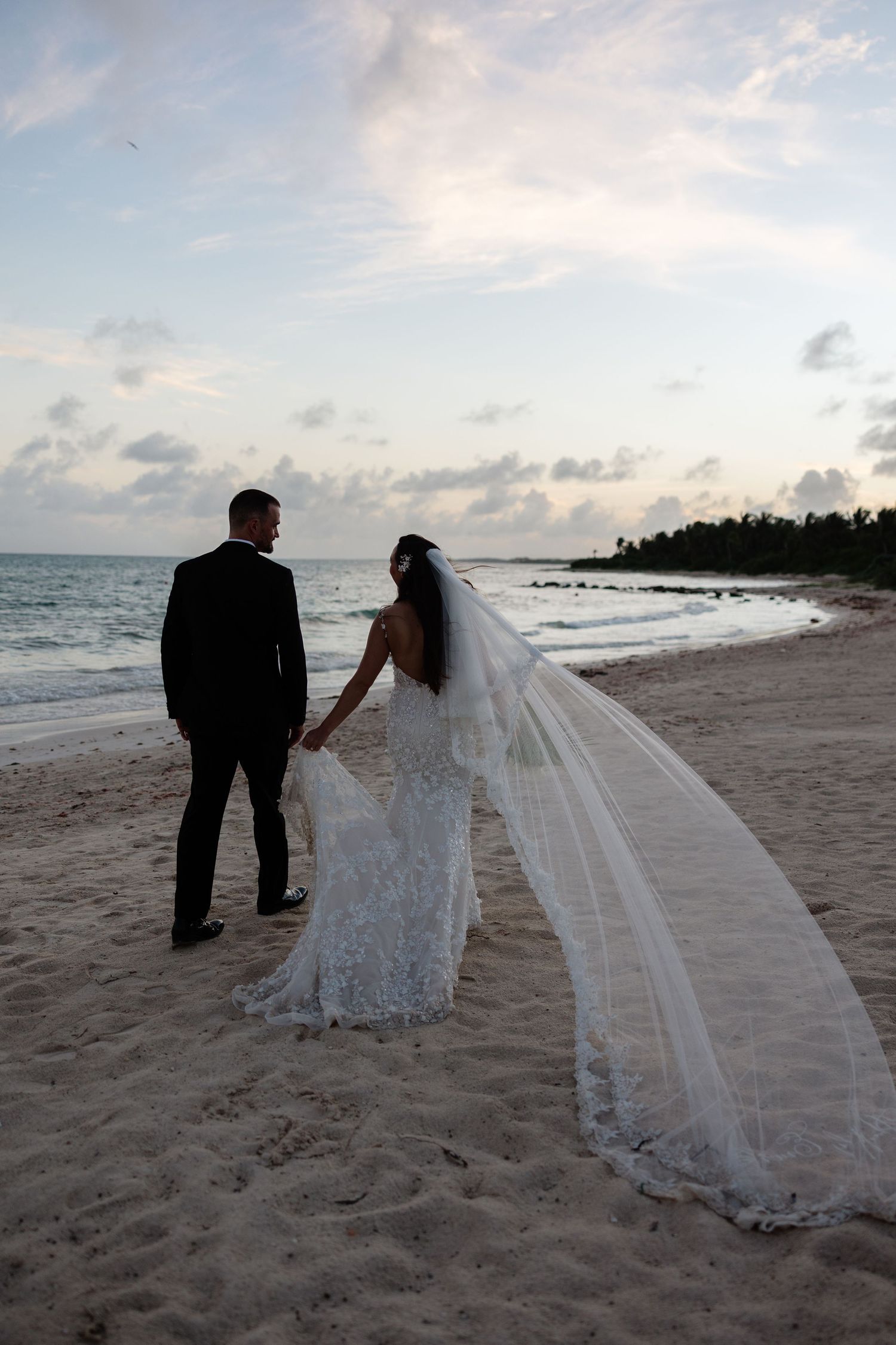 A couple stands on a beach at sunset with a long flowing veil catching the ocean breeze.